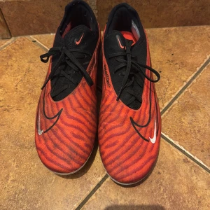 Nike Phantom GX röda fotbollsskor - Nike Phantom GX  - Säljer ett par Nike Phantom GX fotbollsskor i rött och svart med coolt mönster och vita swoosh-detaljer. Skorna har snörning och är designade för gräsplan med dobbar undertill. Perfekta för dig som vill sticka ut på planen! Stolek 41.