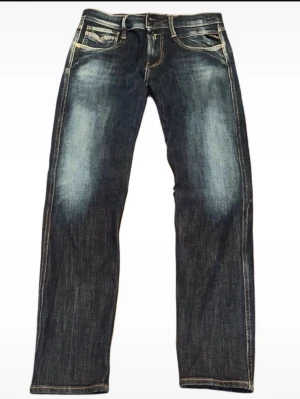 Replay Anbass jeans blå  - Snygga Replay Anbass jeans i mörkblå tvätt med slitningar och kontrastsömmar. Klassisk femficksmodell med raka ben och normal passform. Jeansen har detaljerade bakfickor med broderi och Replay-logga på linningen. Perfekt för dig som gillar stilrena och coola jeans.