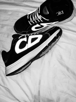 Svarta Dior B30 sneakers med reflexdetaljer - Säljer ett par svarta Dior B30 sneakers med vita och reflekterande detaljer samt tydligt CD-logga på sidan. Skorna har vit sula, meshpaneler och vita snören för en clean look. Perfekta för dig som vill sticka ut med exklusiva och trendiga sneakers.
