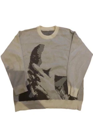  Frank Ocean Knit Sweater - Säljer en beige stickad tröja med ett stort grafiskt motiv i svart och grått framtill. Tröjan har rund halsringning och ribbade muddar vid ärmslut och nederkant. Perfekt för dig som gillar unika prints och en chill vibe.