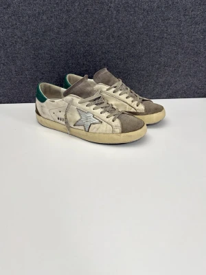 Golden Goose - Säljer ett par Golden Goose sneakers i vitt och grått med gröna detaljer på hälen och den ikoniska stjärnan på sidan. Skorna har snörning, rund tå och är tillverkade i skinn och mocka. Perfekta för dig som gillar en cool och avslappnad stil. Skriv angående frågor!