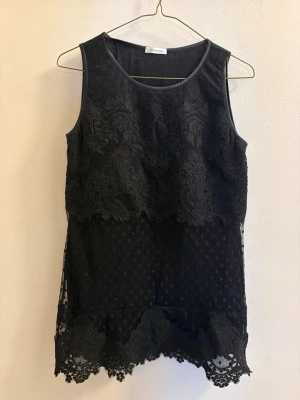 Svart ärmlös spetsblus med prickar - Supersnygg svart ärmlös blus från Ida Sjöstedt med broderade spetsdetaljer och prickigt mönster. Blusen har vackra spetskanter nertill och upptill, samt transparent mesh med prickar som ger en cool och elegant vibe.