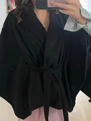 Poncho kappa  - Säljer en svart poncho-kappa med bälte i midjan och bred krage. Jackan har en loose passform och är tillverkad i ett mjukt tyg som känns som ull☺️