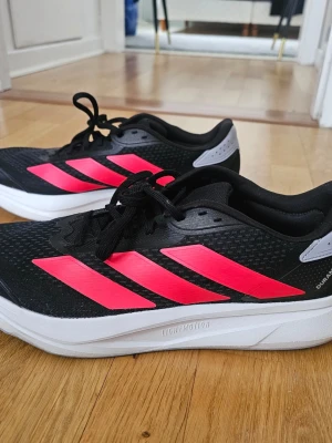 Svarta Adidas sneakers med röda detaljer - Säljer ett par svarta Adidas sneakers med tydliga röda ränder på sidorna och vit sula. Skorna har snörning, mesh-liknande ovandel och grå detaljer vid hälen. Perfekta för träning eller vardagsbruk, riktigt snygga och sportiga. Används bara 1 gång! 46 ⅓ plus 1/3 