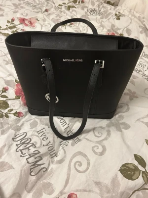 Svart handväska från Michael Kors - Snygg svart handväska från Michael Kors med stilren design och silverfärgade detaljer. Väskan har två långa axelband, ett rymligt huvudfack och ett diskret MK-hänge. Tillverkad i slätt skinn som ger en exklusiv känsla och passar perfekt till alla outfits.