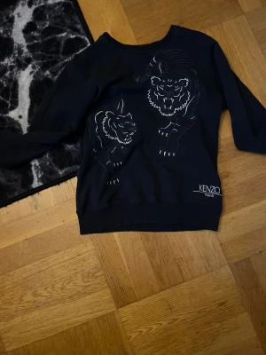Svart sweatshirt med tigerbrodyr från Kenzo - Säljer en svart sweatshirt från Kenzo med två stora broderade tigrar på framsidan och Kenzo Paris-logga nedtill. Tröjan har rund hals, ribbade muddar och är tillverkad i mjuk bomull. Perfekt statement-plagg för dig som gillar unika detaljer.