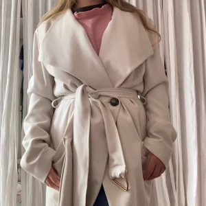 Beige trenchcoat från mbyM 💛 - Snygg beige trenchcoat från mbyM med bred krage och knytbälte i midjan. Jackan har stora fickor, knappar framtill och ett mjukt, glansigt sideninnerfoder. Perfekt för dig som vill ha en stilren och klassisk look. Har en pytteliten lös tråd vid den bruna knappen men syns absolut inte!💛💛💛