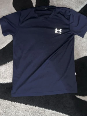 Mörkblå Under Armour t-shirt - Snygg mörkblå t-shirt från Under Armour med klassisk logga på bröstet och liten logga i sidan. Tillverkad i mjukt och lätt polyester som andas, perfekt för träning eller chill. Enkel design med rund hals och korta ärmar. Pris kan diskuteras 