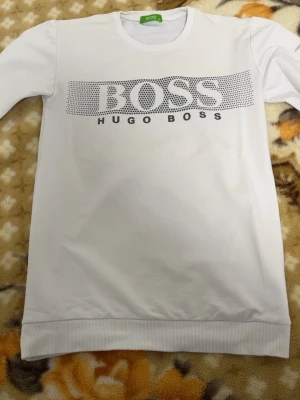 Vit långärmad tröja från Hugo Boss - Snygg vit långärmad tröja från Hugo Boss med stort svart logotryck över bröstet. Tröjan har rund halsringning och ribbade muddar vid ärmslut och nederkant. Perfekt för dig som gillar stilrena och klassiska plagg med tydlig branding.