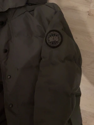 Svart pufferjacka från Canada Goose - Säljer en svart pufferjacka från Canada Goose med huva och patch på ärmen. Jackan har en clean look med knappar framtill och breda quiltade paneler. Perfekt för kalla dagar och riktigt snygg till streetwear-stilen.