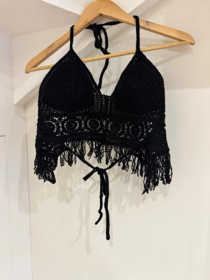 Svart virkad croppad linnetopp - Säljer en svart virkad linnetopp med tunna axelband och knyt i ryggen. Toppen har vadderade kupor, dekorativa virkade mönster och fransar längs nederkanten. Perfekt för sommarens alla äventyr och festivaler.