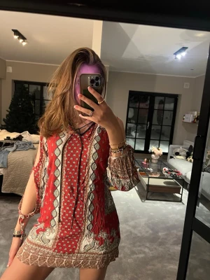 Röd boho-inspirerad kortklänning - Säljer en röd kortklänning med boho-vibbar och mönster i beige och vitt. Klänningen har v-ringning, långa ärmar med öppna slitsar och knytband samt ett luftigt, flowy material. Perfekt för dig som gillar färg och mönster!