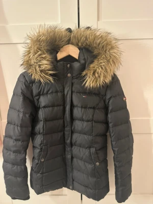  pufferjacka  - Säljer en svart pufferjacka från Hilfiger Denim med stor, fluffig fuskpäls på huvan. Jackan har quiltad design, dragkedja och knappar framtill samt två fickor med tryckknappar. Perfekt för kalla dagar och riktigt snygg till vintern.