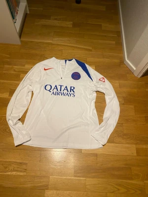 PSG X Nike Tracksuit 25/26 Player Version Helt Ny!!! - Säljer ett helt nytt träningsset från Paris Saint-Germain, säsongen 2025/2026. Setet består av en långärmad vit träningströja och svarta träningsbyxor – båda med PSG:s officiella klubbmärke och Nike-logga i rött. Tröjan har quarter-zip och tryckt sponsor “Qatar Airways” på bröstet. Byxorna har elastisk midja med snörning, blå sidodetalj och matchande emblem.