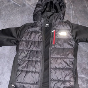 Svart pufferjacka från The North Face - Säljer en svart pufferjacka från The North Face med huva och quiltad framsida. Jackan har en röd dragkedja på bröstfickan och reflekterande logga. Materialet är polyester och jackan har långa ärmar. Perfekt för kyliga dagar.