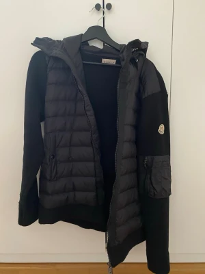 Svart Moncler Cardigan med Luva - Säljer en svart pufferjacka från Moncler med stickade armar och quiltad kropp. Storlek M. Jackan har luva, dragkedja framtill och en ficka med dragkedja på armen. Perfekt för kyliga dagar och riktigt snygg streetstil.