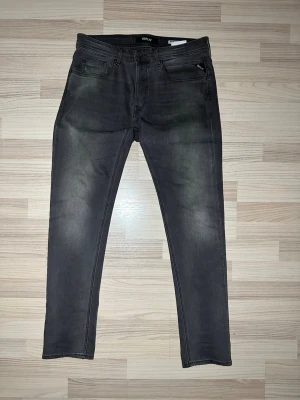 Replay Anbass mörkgrå jeans - Säljer ett par Replay Anbass jeans i mörkgrå tvätt med snygga slitningar och klassisk femficksdesign. Jeansen har smal passform och raka ben, med Replay-logga på bakfickan och läderpatch i midjan. Perfekt för dig som gillar stilrena och moderna jeans.