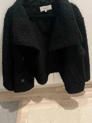 Svart teddyjacka från Bubbleroom - Säljer en svart teddyjacka från Bubbleroom med bred krage och dubbelknäppning framtill. Jackan har en fluffig och mjuk yta, långärmad design och en loose passform som ger en chill vibe. Perfekt för dig som gillar stilrena och mysiga plagg.