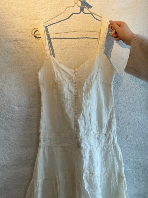 Vit somrig kortklänning med knappar - Säljer en luftig vit kortklänning med breda axelband och dekorativa knappar framtill. Klänningen har en lätt rynkad kjol och är perfekt för varma dagar. Tillverkad i ett tunt, mjukt material som ger en avslappnad vibe.