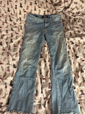 Replay FAABY ljusblå croppade jeans. 3/4 passform - Säljer ett par ljusblå Replay FAABY jeans med croppad flare fit och midwaist. Klassisk femficksmodell med snygga slitningar och rå kant nedtill. Broderad R-detalj på fickan och tydlig Replay-logga i linningen. Perfekta för dig som gillar en relaxed men trendig look.