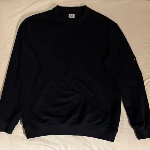 Mörkblå sweatshirt från C.P. Company - Hej! Säljer en tvärfet mörkblå sweatshirt från C.P. Company med klassisk rund hals och lång ärm. Tröjan har en ikonisk linsdetalj på ena ärmen och en liten ficka med dragkedja. Tillverkad i mjuk bomull för en skön och avslappnad look. Skriv om du har några frågor eller vid intresse. 