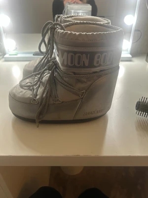 Silvriga Moon Boots med snörning - Säljer ett par klassiska silvriga Moon Boots med snörning och rund tå. Skorna har en hög och vadderad design som ger en futuristisk vibe och är perfekta för vintern. De är tillverkade i syntetmaterial och har en platt sula för extra komfort. Pris kan diskuteras