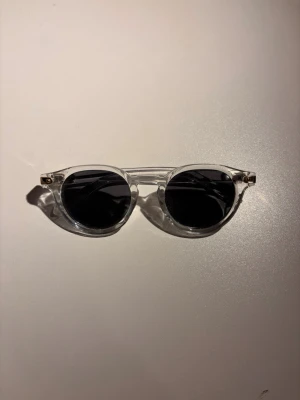 Solglasögon  - Snygga solglasögon med transparent båge och runda svarta glas. Perfekt accessoar för att lyfta din stil och skydda ögonen mot solen. Klassisk och trendig look som passar till det mesta.
