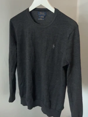 Polo Ralph Lauren tröja  - Säljer en mörkgrå långärmad tröja från Polo Ralph Lauren i slim fit. Tröjan är tillverkad i tvättbar merinoull och har en klassisk rund halsringning samt den ikoniska broderade loggan på bröstet.