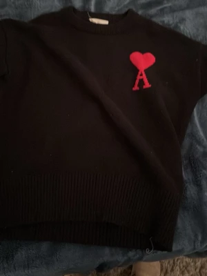 Svart stickad tröja från Ami Paris - PASSAR 160 Säljer en svart stickad tröja från Ami Paris med ikonisk röd hjärtlogga och bokstaven A på bröstet. Tröjan har rund hals, ribbade muddar och lång ärm. Perfekt för dig som vill ha en clean men ändå statement look.