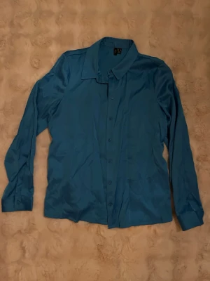 Blå skjorta från vero moda i satinlook - Snygg blå skjorta från vero moda i satinliknande material. Klassisk krage, långärmad och med knappar hela vägen framtill. Perfekt för dig som vill ha en stilren och enkel look med lite extra glans.