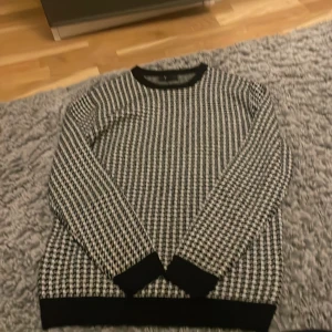 Svartvit rutig tröja från SMOG - Säljer en svartvit tröja från SMOG i regular fit med klassiskt rutigt mönster. Tröjan har svarta muddar vid ärmslut, nederkant och halsringning. Perfekt till höst och vinter för en clean och stilren look.