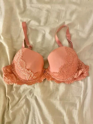 Rosa spets-bh med rosett, 70C💗 - Söt bh i persika rosa med fina spetsdetaljer längs kanterna. Dekorerad med en liten rosett framtill. Justerbara axelband för bästa passform. 