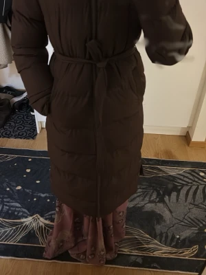 Brun lång pufferjacka med bälte - Säljer en lång, mörkbrun pufferjacka med bälte i midjan och dragkedja framtill. Jackan har långa ärmar och en clean, stilren look som passar perfekt när det är kallt ute. Fylld med vaddering för extra värme och har praktiska fickor på sidorna.