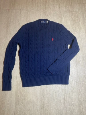 Marinblå kabelstickad tröja Polo Ralph Lauren - Kabelstickad marinblå tröja från Polo Ralph Lauren med klassisk röd logga broderad på bröstet. Tröjan har rund halsringning, ribbade muddar och är långärmad. Perfekt för dig som gillar stilrena och tidlösa plagg. Liten i storlek och även helt oanvänd, endast att lappen lostnat.