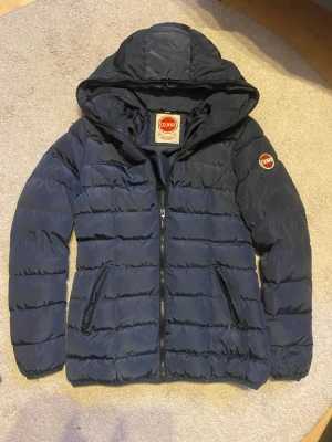 Colmar Originals jacka – storlek M - Snygg och varm Colmar Originals jacka i mörkblå färg. Vadderad modell med huva, dragkedja framtill och två fickor med dragkedja. Klassisk Colmar-logga på ärmen.  Jackan är i bra skick, sparsamt använd och utan hål, fläckar eller andra större slitage. Perfekt för höst och vinter.  📏 Storlek: M 👕 Passform: Normal 🏷️ Märke: Colmar Originals 📦 Skick: Bra  Hör gärna av dig vid frågor! 😊