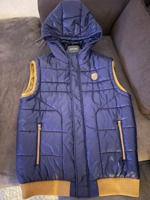 Marinblå dunväst från Antony Morato - Snygg marinblå dunväst från Antony Morato med huva och guldfärgade detaljer. Västen har två fickor med dragkedja, ribbade muddar och ett coolt Morato Gold-märke på bröstet. Perfekt för lager på lager och streetwear-vibe.