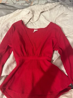 Röd omlottopp från Nelly - Säljer en snygg röd omlottopp från Nelly i storlek S. Toppen har lång ärm, v-ringning och ett band i midjan som knyts baktill. Materialet är ribbat och ger en cool struktur. Perfekt för dig som vill ha något extra till outfiten.
