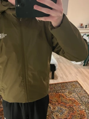 Olivgrön vindjacka från The North Face - Säljer en olivgrön vindjacka från The North Face i storlek XS. Jackan har huva, dragkedja framtill och diskret logga på bröstet. Tillverkad i slitstark polyester och har en clean, minimalistisk look som passar perfekt till streetwear.