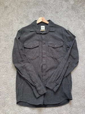 Mörkgrå skjorta från Dobber - Stilren mörkgrå skjorta från Dobber med klassisk krage och två bröstfickor med lock. Skjortan har långa ärmar och knäppning framtill. Perfekt för dig som gillar en enkel men snygg look.                      Bara att höra av sig vid eventuella frågor!