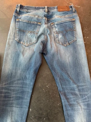 Tiger Of Sweden Jeans - Säljer ett par blå Tiger Of Sweden jeans med snygga slitningar och en trasig detalj på vänster knä. Jeansen har klassisk femficksdesign, normal midja och raka ben. Perfekta för dig som gillar en avslappnad och trendig look.