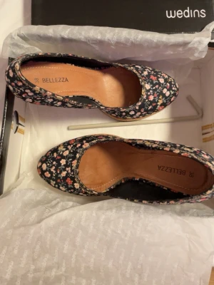 Blommiga pumps från Bellezza - Snygga pumps från Bellezza med svart bas och blommigt mönster i rosa, vitt och grönt. Skorna har spetsig tå, hög klack och plattform i korkliknande material. Perfekta för dig som vill sticka ut med en färgglad och trendig look.