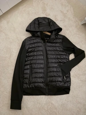 Svart dunjacka med huva från Khujo - Snygg svart dunjacka från Khujo med quiltad framsida och huva. Jackan har dragkedja framtill, två fickor med dragkedja och släta svarta ärmar. Perfekt för kalla dagar när du vill hålla stilen.