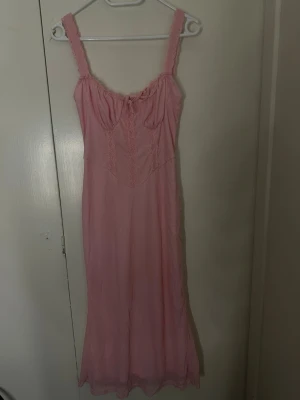 Rosa klänning med spetsdetaljer - Romantisk ljusrosa långklänning med breda axelband och fina spetsdetaljer framtill. Klänningen har en feminin siluett och mjukt fallande tyg, perfekt för dig som gillar en söt och klassisk stil.