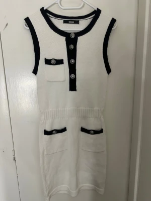 Vit stickad ärmlös klänning  - Vit stickad klänning från bikbok med svarta kontrastkanter, dekorativa knappar framtill och tre fickor. Ärmlös modell med rund hals och ribbad midja. Perfekt för en trendig och chic look.