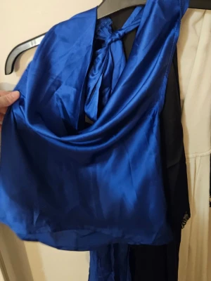 Blå ärmlös blus med knytband - Snygg blå blus i glansigt material med dekorativt långt knytband framtill. Blusen är ärmlös och har en elegant, lös passform som ger en lyxig känsla. Perfekt för att lyfta vilken outfit som helst.
