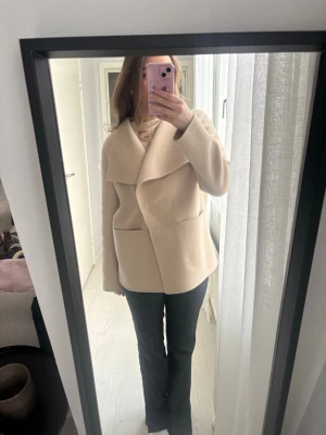 Beige omlottkappa  - Säljer en snygg beige kappa med omlottknäppning och stor, bred krage. Kappan har två fickor framtill och går att knäppa eller ha öppen. På insidan av jackan finns en liten fläck av ett par blå jeans som färgat av sig. Denna syns inte varken om du har kappan stängd eller öppen, men har inte vågat tvätta då kemtvätt rekommenderas!