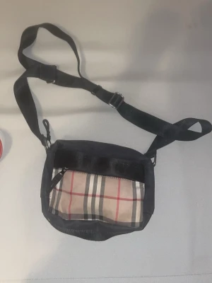 Rutig axelväska från Burberry - Snygg axelväska från Burberry med klassiskt rutigt mönster i beige, svart och rött. Väskan har justerbar axelrem och praktiskt innerfack. Perfekt för att bära dina viktigaste grejer med stil.