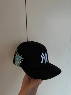 Svart New York Yankees keps ”fitted” - Snygg svart keps från New Era med New York Yankees-logga i vitt framtill och broderad Frihetsgudinna i turkos på sidan. Platt skärm och klassisk snapback-modell. Perfekt för dig som gillar streetwear och vill sticka ut med detaljer.  Storlek 7 3/8 eller 58,7.