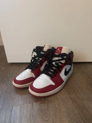 Nike Air Jordan 1 Mid Chicago - Säljer ett par Nike Air Jordan 1 Mid Chicago med klassisk färgkombination i vitt, rött och svart. Skorna har svart snörning, rund tå och den ikoniska swoosh-loggan på sidan. Tillverkade i skinn med perforerad tåbox och vadderad krage för extra komfort.