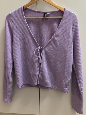 Ljuslila ribbad topp från H&M Divided - Säljer en ljuslila ribbad topp från H&M Divided. Toppen har lång ärm, croppad passform och v-ringning med knyt framtill. Perfekt att styla med höga jeans eller kjol för en trendig look.